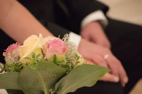 Detailaufnahme eines Hochzeitspaars, sitzend und Händchen haltend, mit Blumengebinde im Vordergrund